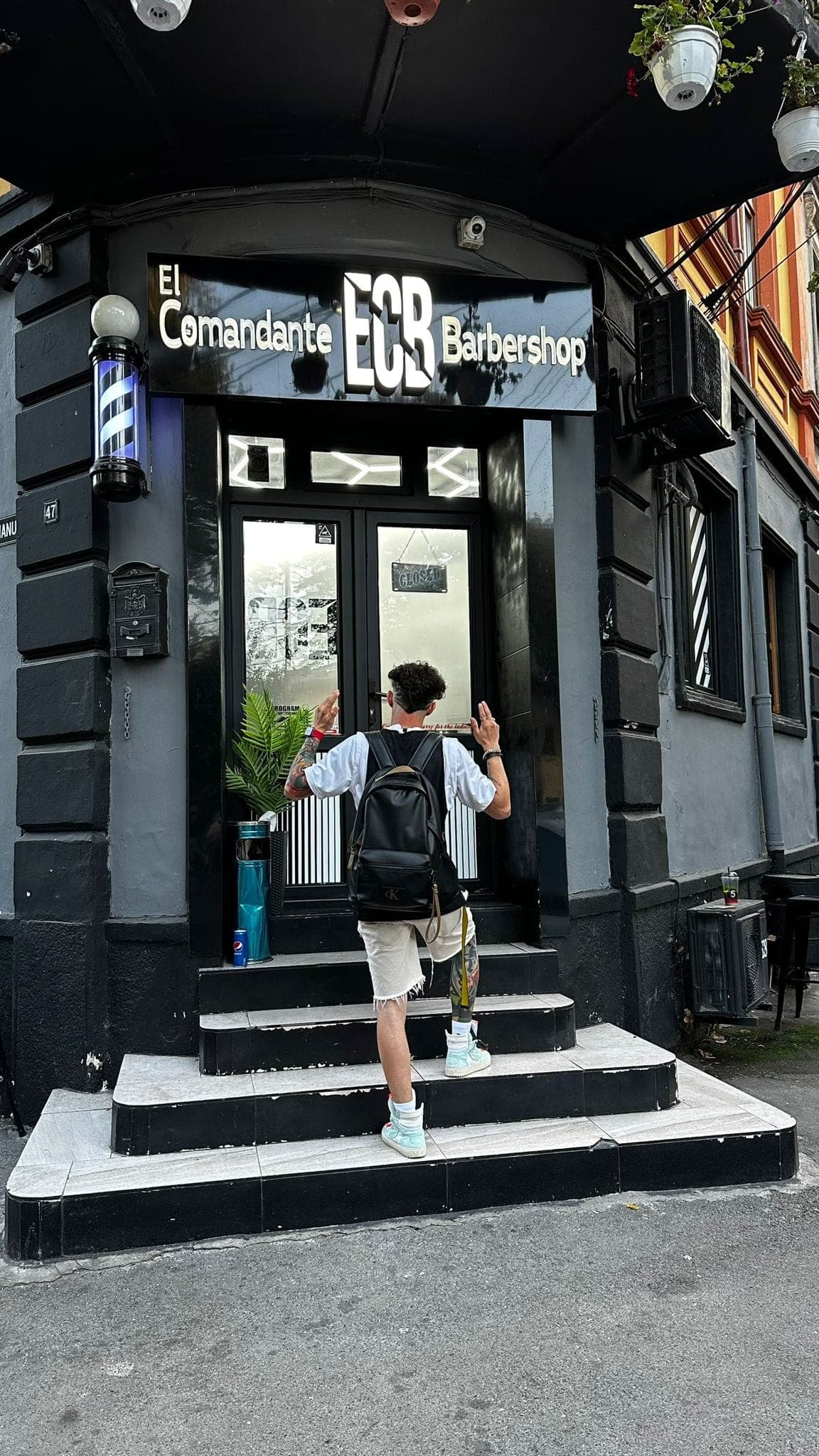 Tânăr intrând în El Comandante Barbershop — vedere largă cu signajul complet și stâlpul de frizer